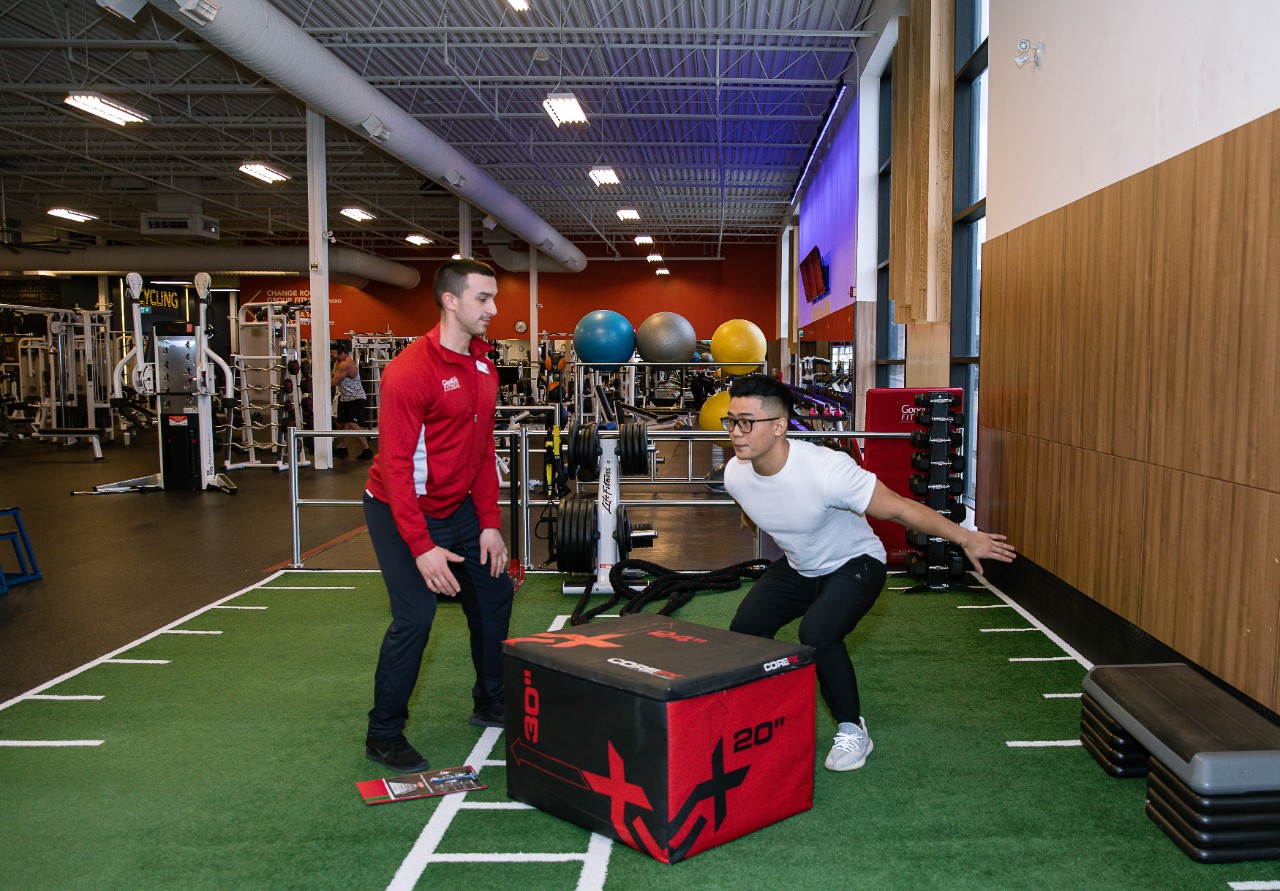 A Personal Trainer assisting a Member with box jumps in the turf area at a GoodLife Fitness Club